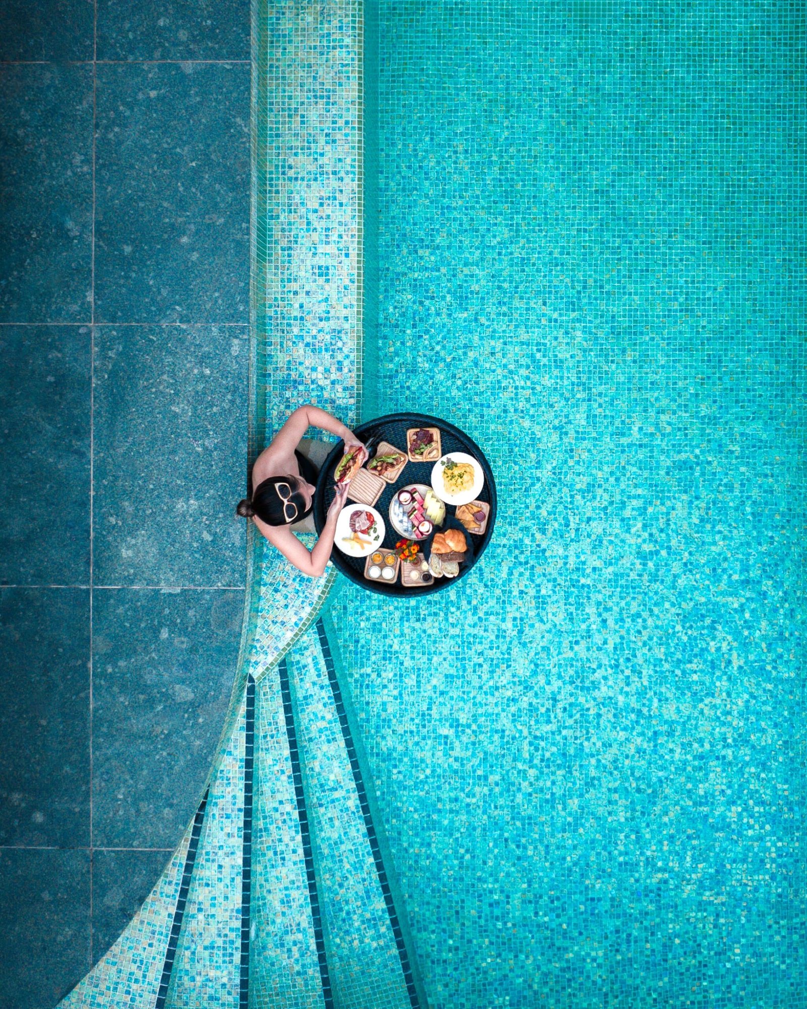 Top-down drone shot of a woman in a pool, enjoying a floating breakfast.